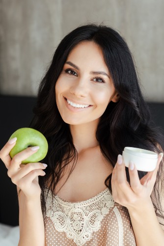 Smiling woman holding a green apple in one hand and a jar of face cream in the other
