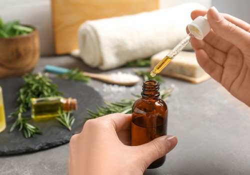 Hands lifting a dropper of golden oil from an amber bottle beside green herbs and a rolled towel