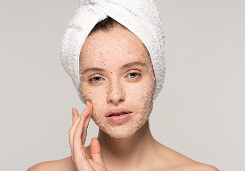 Woman with a towel around her hair touching the exfoliating scrub on her cheek
