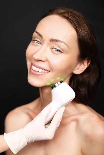 Gloved hand holding a white skincare device with green lights against a woman's jawline