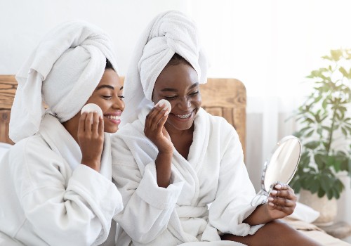 Two women in bathrobes using cotton pads to cleanse their faces beside a handheld mirror