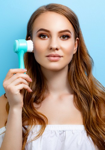 Woman holding a turquoise electric cleansing brush beside her cheek against a blue background