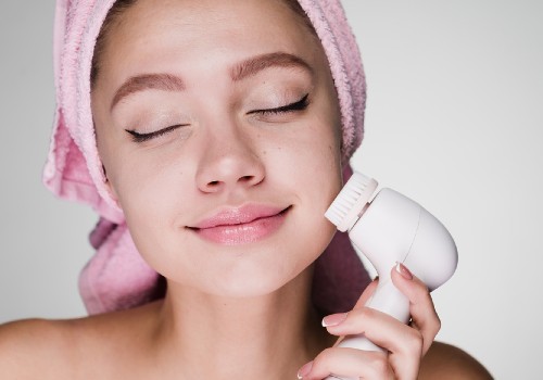 Woman with her hair in a pink towel holding an electric cleansing brush against her cheek