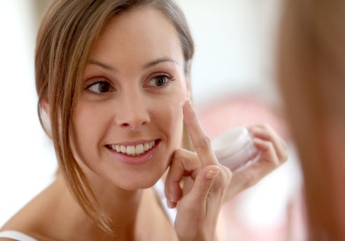 Smiling woman dabbing moisturizer onto her cheek while holding an open jar