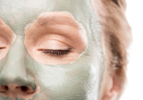 Close-up of a woman's face covered with a pale green clay mask, with the eye area left clear