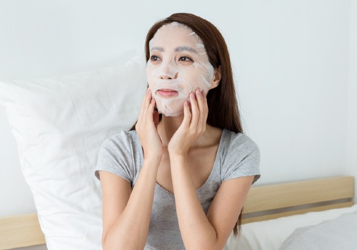 Woman sitting on a bed and smoothing a white sheet mask against her cheeks