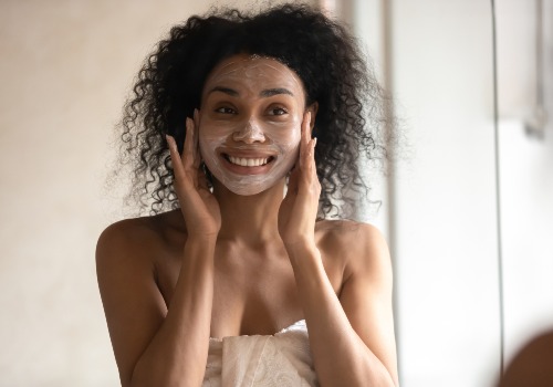 Woman massaging a facial mask onto both cheeks while looking in a mirror