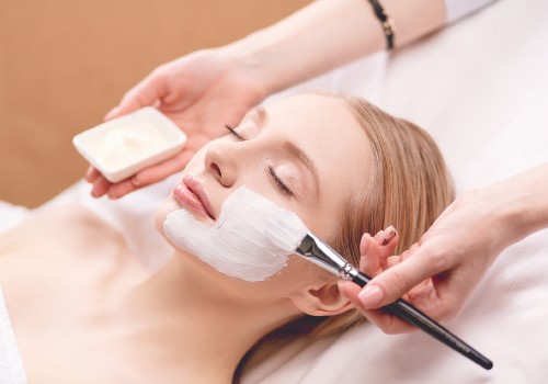 Practitioner applying a white facial mask to a reclining woman's cheek with a flat brush