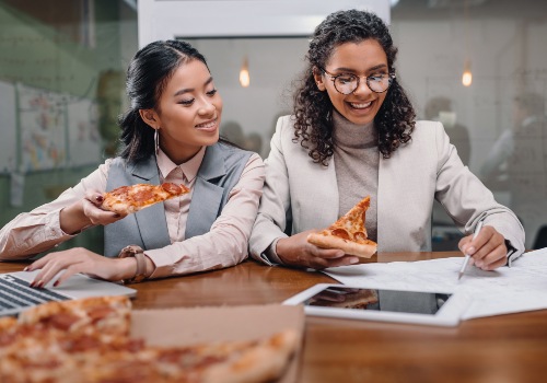 Two women eating pizza at a worktable with a laptop and paperwork