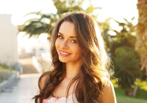 Smiling woman with long wavy hair standing outdoors beside a white wall and plants