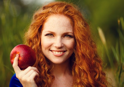 Red-haired woman with freckles holding a red apple beside her face outdoors