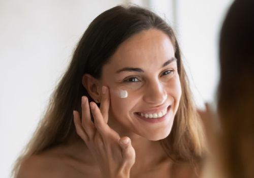 Woman dabbing eye cream onto the skin beneath her eye while looking in a mirror