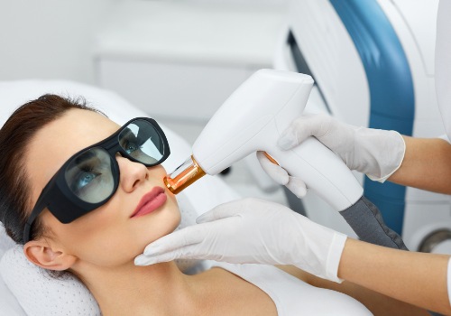 Practitioner holding a treatment device beside a woman's upper lip while she wears protective goggles