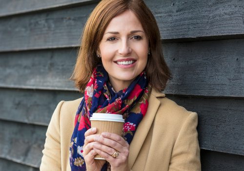 Smiling woman in a colorful scarf holding a takeaway cup against a dark wooden wall