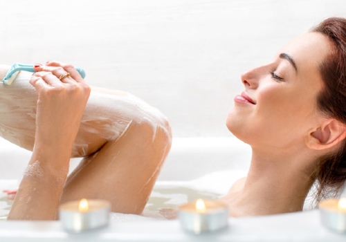 Woman shaving a cream-covered leg while reclining in a bathtub beside lit candles