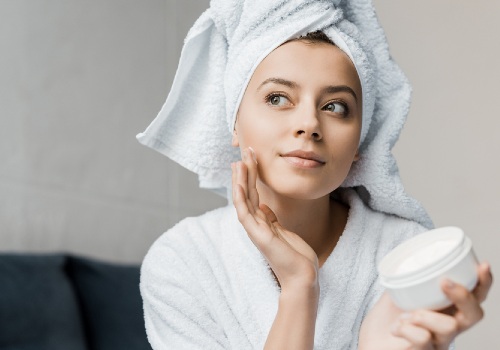Woman with her hair wrapped in a towel touching her cheek while holding an open moisturizer jar