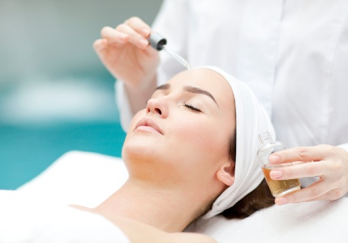 Practitioner holding a glass dropper above a reclining woman's forehead during a facial treatment