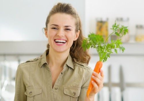 Smiling woman holding a fresh carrot with leafy green tops in a kitchen
