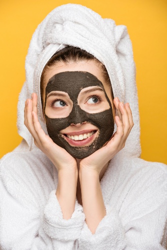 Woman in a bathrobe wearing a black facial mask and holding her hands against her cheeks