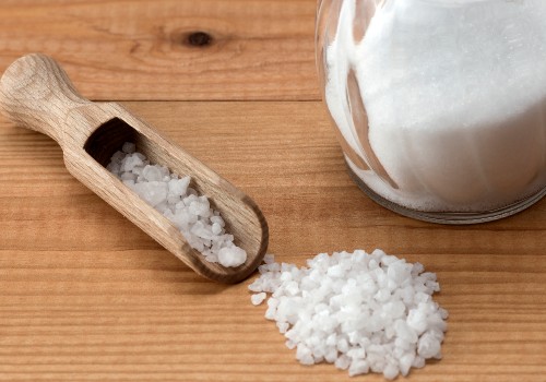 Wooden scoop of coarse salt beside a jar of granulated sugar on a wooden surface