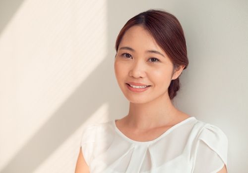Smiling woman with her hair tied back wearing a white blouse beside a sunlit wall