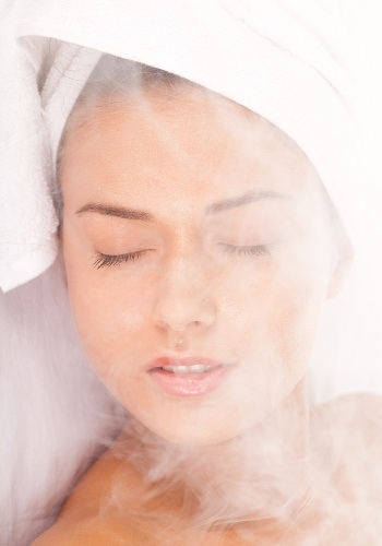 Woman with a towel draped over her head holding her face above rising steam