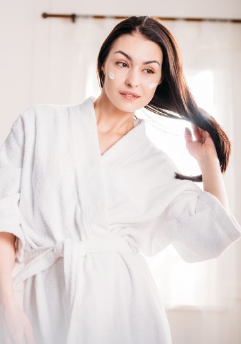 Woman in a white bathrobe standing in a bright room with one hand raised to her hair