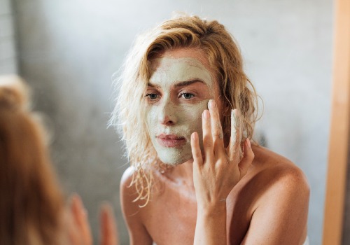 Woman smoothing a pale green face mask onto her cheek while looking in a mirror