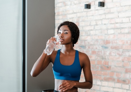 Woman in blue sportswear drinking water from a clear bottle beside a brick wall