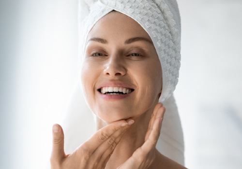 Smiling woman with a towel around her hair massaging moisturizer along her chin and neck